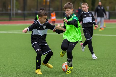 Zwei Kinder im Fußballtrikot kämpfen um den Ball auf einem Sportplatz.