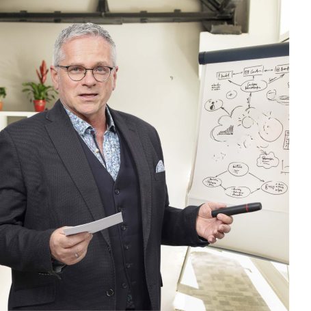 Mann mit Brille in Anzug hält ein Blatt und einen Stift, steht vor einer Präsentation.