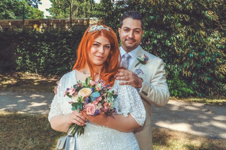 Braut und Bräutigam posieren lächelnd im Freien mit Blumenstrauß.
