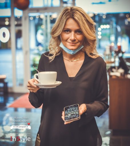 Frau mit blonden Haaren, hält eine Tasse Kaffee und ein Smartphone, lächelt freundlich.