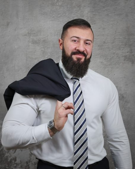 Mann mit Bart, Anzugjacke auf der Schulter und gestreifter Krawatte vor neutralem Hintergrund.