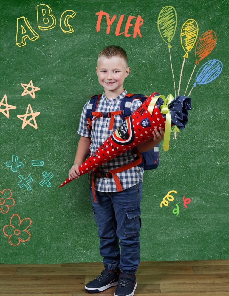 Einschulungsshooting Adler Photographie Ein Junge mit Rucksack hält einen bunten Schultütenhalter vor einer grünen Tafel.