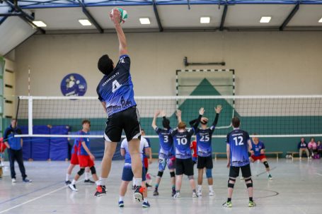 Spieler springt zum Aufschlag beim Volleyball, Teams in blauen und roten Trikots.
