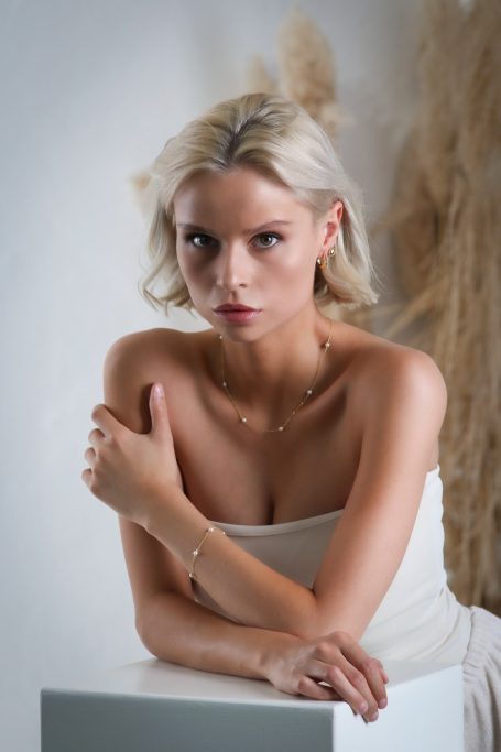 Junge Frau mit blonden Haaren, eleganten Kleid und nachdenklichem Ausdruck vor hellem Hintergrund.