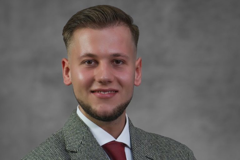 Mann mit kurzem Haar, Bart und grauem Anzug, lächelt vor einem neutralen Hintergrund.