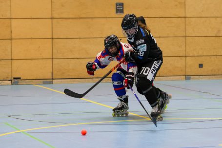 Zwei Spielerinnen beim Rollhockey, eine mit Puck, in einer Sporthalle.