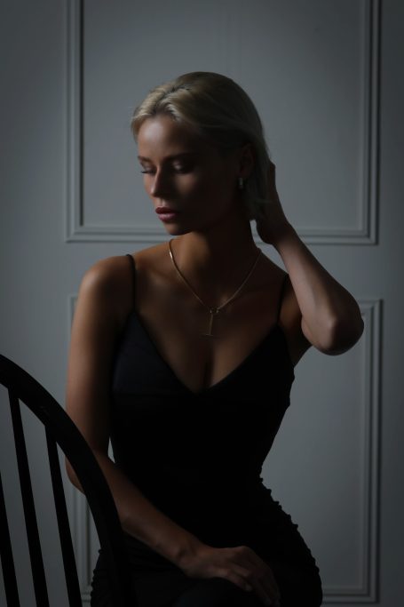 Junge Frau in schwarzem Kleid sitzt nachdenklich vor einer hellen Wand.
