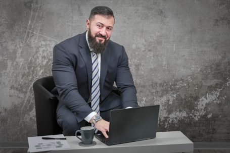 Mann mit Bart in Anzug sitzt am Schreibtisch und arbeitet am Laptop. Tasse daneben.