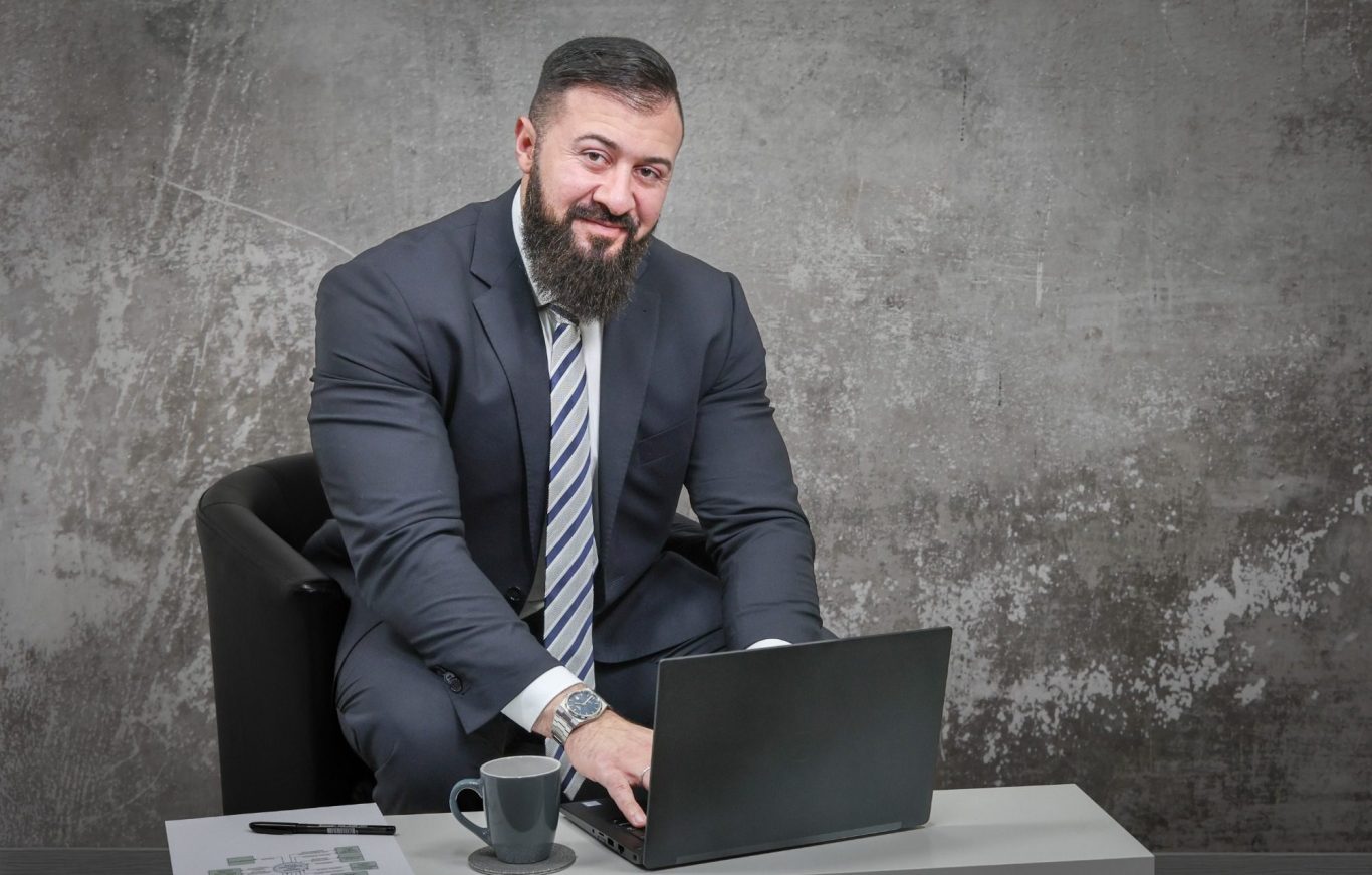 Business shooting krefeld Mann im Anzug sitzt an einem Tisch mit Laptop und Kaffeetasse, lächelt freundlich.