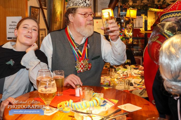 Mann mit Bart und Krone hebt ein Glas, Frau sitzt neben ihm, beide in geselliger Runde.