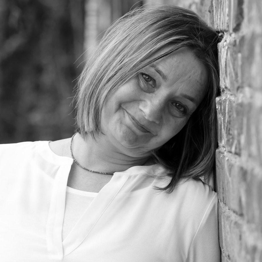 Fotoshooting Adler Photographie Krefeld Frau mit kurzem, blondem Haar, lächelnd vor einer Backsteinmauer.