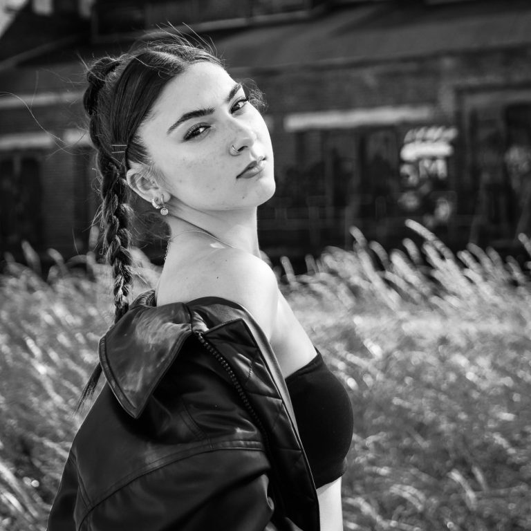 Fotoshooting Krefeld Adler Photographie Junge Frau mit Zöpfen, trägt ein schwarzes Top, steht in einem Grasfeld.