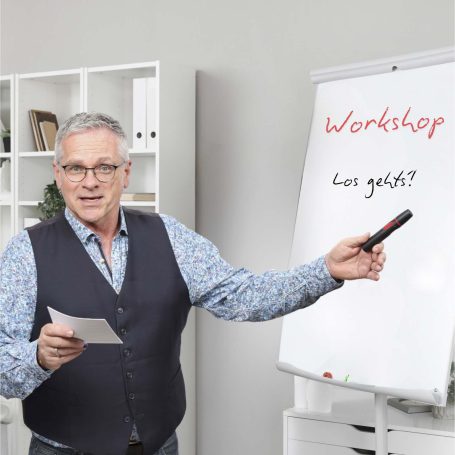 Mann in Weste hält Notizen und zeigt auf ein Flipchart mit "Workshop: wie geht's?".