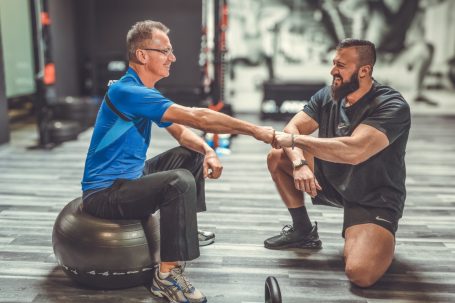 Zwei Männer trainieren gemeinsam in einem Fitnessstudio, einer sitzt auf einem Gymnastikball.