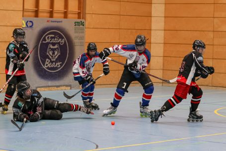 Fünf Inline-Hockeyspieler in Aktion, einer fällt zu Boden, eine Kugel rollt im Vordergrund.