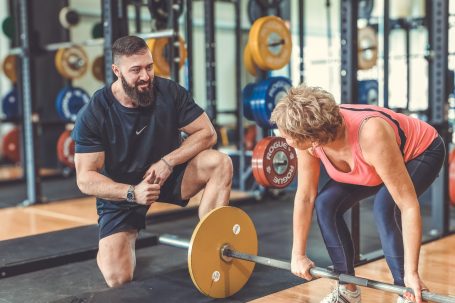 Ein Trainer unterstützt eine ältere Frau beim Gewichtheben in einem Fitnessstudio.