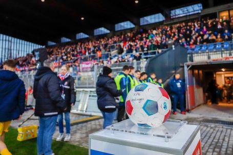 Voller Stadion mit Zuschauern, einer Gruppe am Eingang und einem Fußball in der Vordergrund.