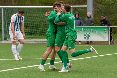 Drei jubelnde Fußballspieler in grünen Trikots auf dem Platz, ein Spieler im Hintergrund.