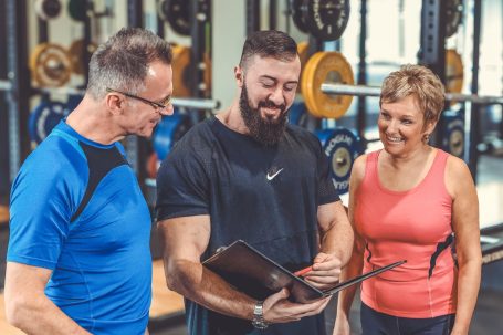 Drei Personen im Fitnessstudio besprechen einen Trainingsplan. Gewichte im Hintergrund sichtbar.