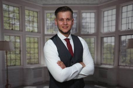 Junger Mann in Anzug mit verschränkten Armen vor großen Fenstern.