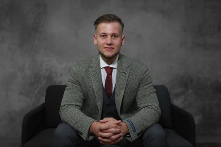 Ein junger Mann im grauen Anzug sitzt lächelnd auf einem schwarzen Sofa.