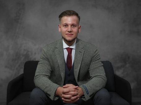 Businessshooting krefeld Adler Photographie Mann in einem Anzug sitzt auf einem Sofa vor einem grauen Hintergrund.